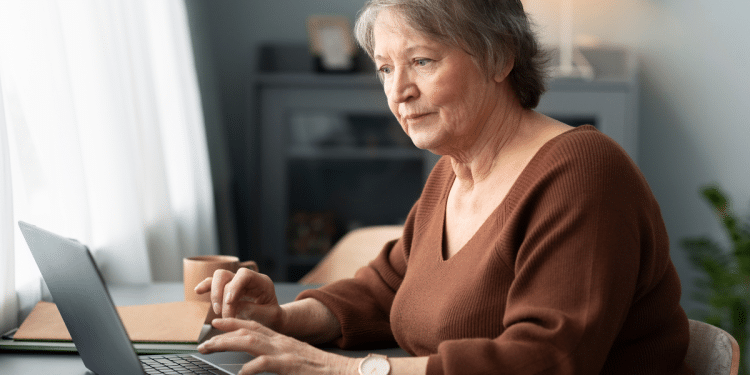Mulher idosa com suéter marrom trabalhando e navegando em notebook próximo à janela iluminada