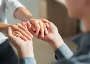 Duas pessoas segurando as mãos demonstrando apoio, confiança e empatia em ambiente interno.