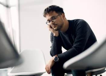 Homem com óculos e suéter preto olhando pensativo sentado em cadeira branca em ambiente claro.