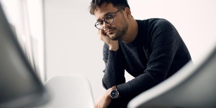 Homem com óculos e suéter preto olhando pensativo sentado em cadeira branca em ambiente claro.