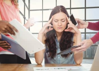 Mulher com expressão ansiosa cercada por várias pessoas oferecendo documentos e dispositivos eletrônicos em escritório moderno.