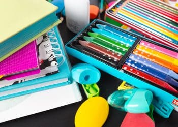 Cadernos, lápis de cor, giz de cera e outros materiais escolares coloridos organizados sobre mesa preta.