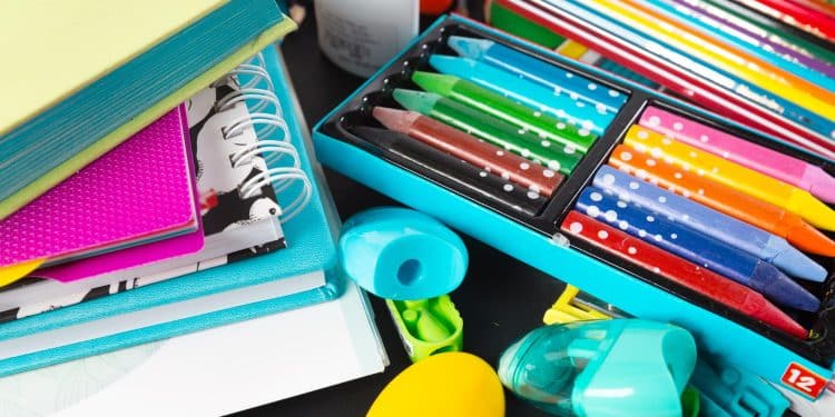 Cadernos, lápis de cor, giz de cera e outros materiais escolares coloridos organizados sobre mesa preta.