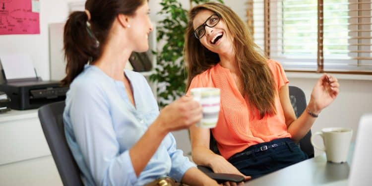 Duas mulheres conversando e sorrindo enquanto tomam café em ambiente de escritório iluminado