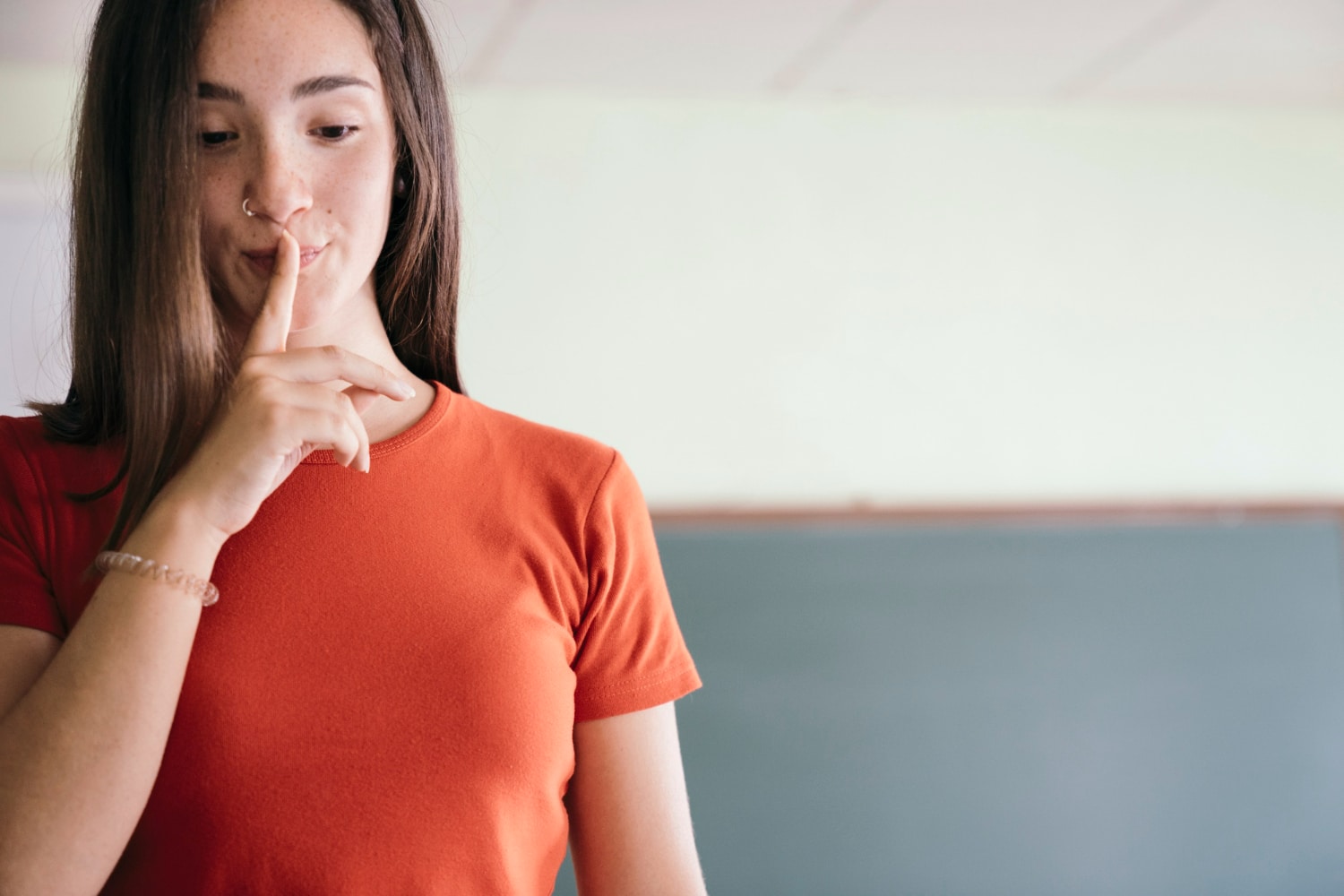Mulher jovem com camiseta laranja fazendo sinal de silêncio com o dedo indicador na frente dos lábios.