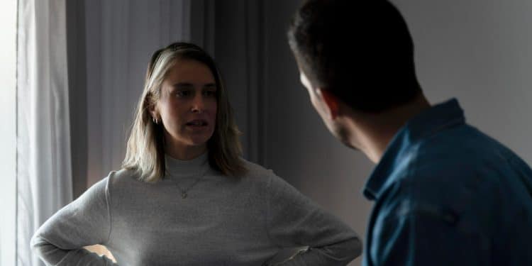Mulher com blusa cinza conversando seriamente com homem de camiseta azul dentro de casa.
