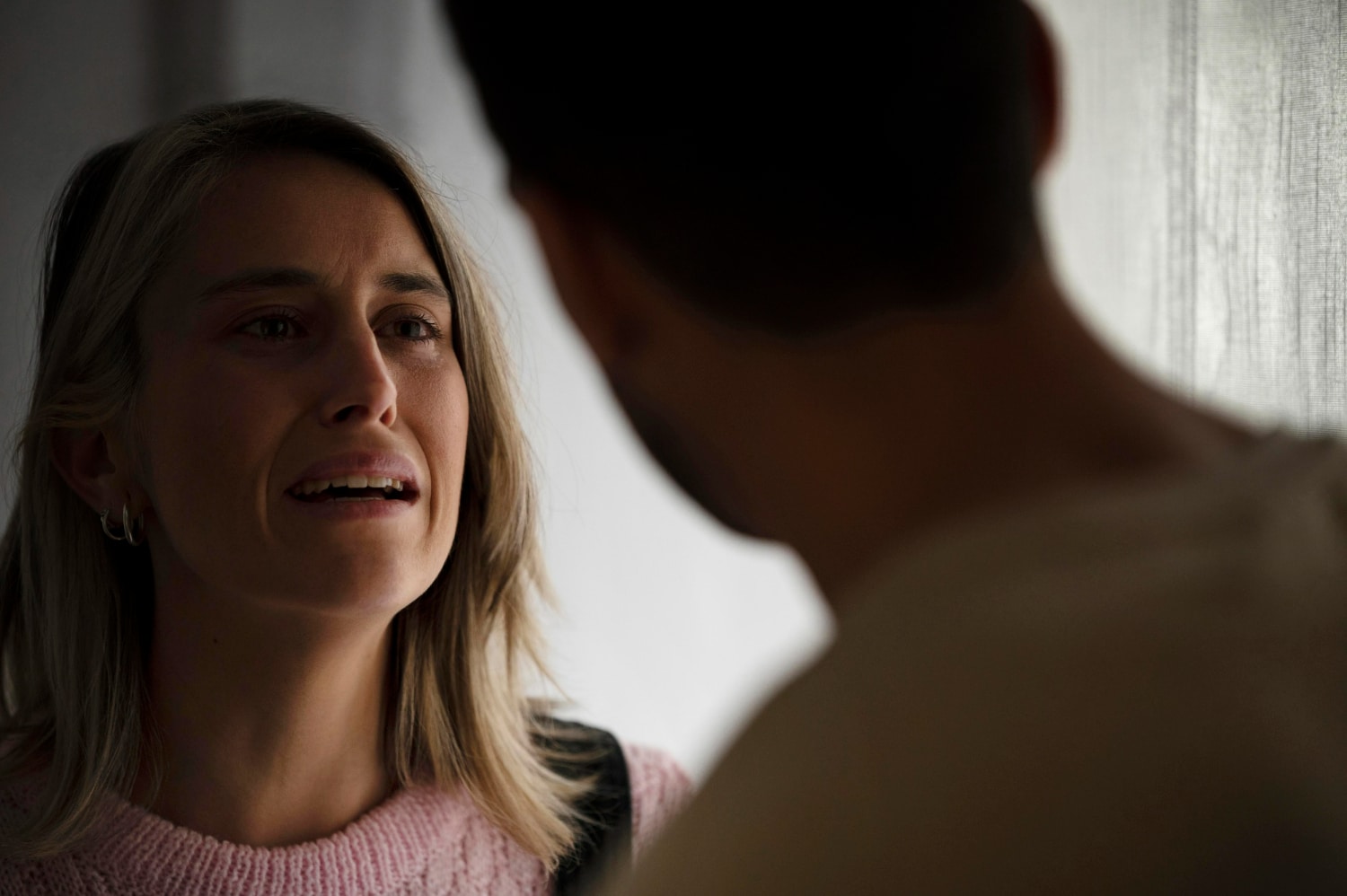 Mulher com blusa cinza conversando seriamente com homem de camiseta azul dentro de casa.