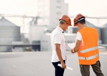 Dois engenheiros com capacete de segurança analisando documento em área industrial.