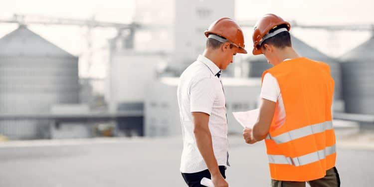 Dois engenheiros com capacete de segurança analisando documento em área industrial.