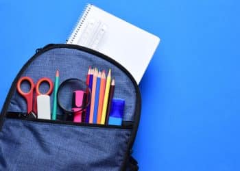 Mochila azul aberta com material escolar incluindo cadernos, lápis de cor, tesoura e régua sobre fundo azul