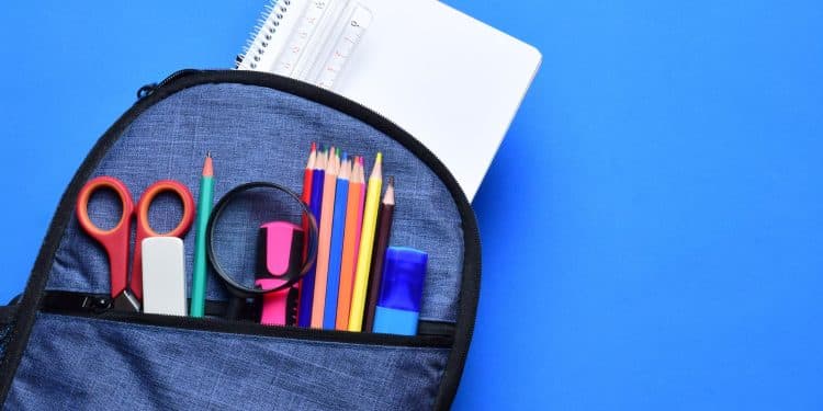 Mochila azul aberta com material escolar incluindo cadernos, lápis de cor, tesoura e régua sobre fundo azul