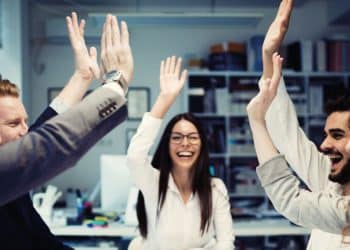 Grupo de colegas de trabalho sorrindo e celebrando juntos no escritório, exemplificando a conexão social como estratégia de felicidade profissional.