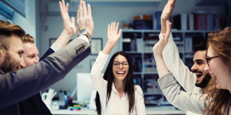 Grupo de colegas de trabalho sorrindo e celebrando juntos no escritório, exemplificando a conexão social como estratégia de felicidade profissional.