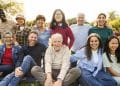 Grande grupo multiétnico reunindo diversas gerações, de idosos a jovens, sorrindo e sentados juntos em uma área verde ao ar livre.