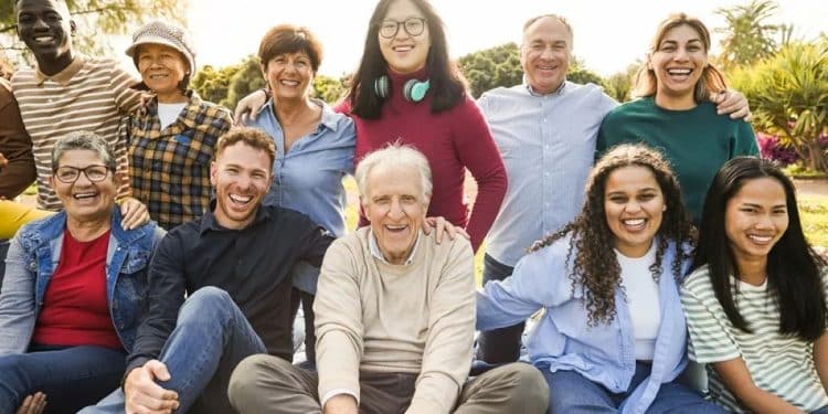 Grande grupo multiétnico reunindo diversas gerações, de idosos a jovens, sorrindo e sentados juntos em uma área verde ao ar livre.
