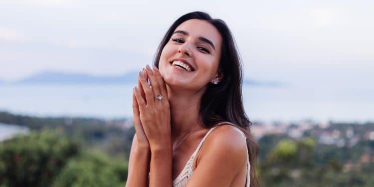 Mulher jovem sorridente com cabelo escuro, usando vestido branco e anel de prata nas mãos ao ar livre Legenda: Jovem feliz exibindo seu anel de prata em ambiente ao ar livre com paisagem desfocada ao fundo.
