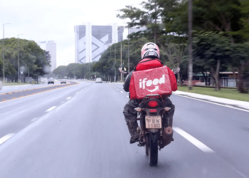 Motoboy parceiro do iFood pilotando motocicleta em avenida urbana com bag de entrega vermelha característica da plataforma.