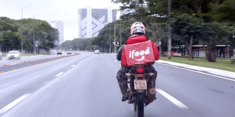 Motoboy parceiro do iFood pilotando motocicleta em avenida urbana com bag de entrega vermelha característica da plataforma.