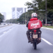 Motoboy parceiro do iFood pilotando motocicleta em avenida urbana com bag de entrega vermelha característica da plataforma.