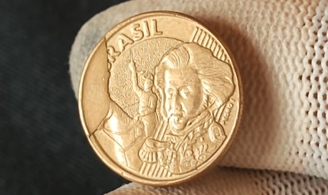 Close-up de uma moeda brasileira de 10 centavos com um erro visível de rachadura (cunho quebrado) no campo esquerdo. O defeito é típico do ano 2001 e a torna um item raro de colecionador, valendo até R$ 400.