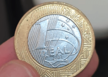 Close-up de uma moeda de 1 Real com defeito, sendo segurada por um numismata. A peça de 2021 com erro de reverso pode ser vendida por até R$ 160 no mercado de colecionadores.