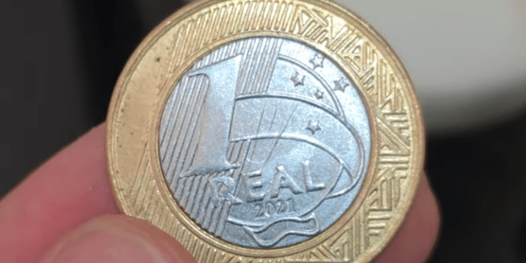 Close-up de uma moeda de 1 Real com defeito, sendo segurada por um numismata. A peça de 2021 com erro de reverso pode ser vendida por até R$ 160 no mercado de colecionadores.