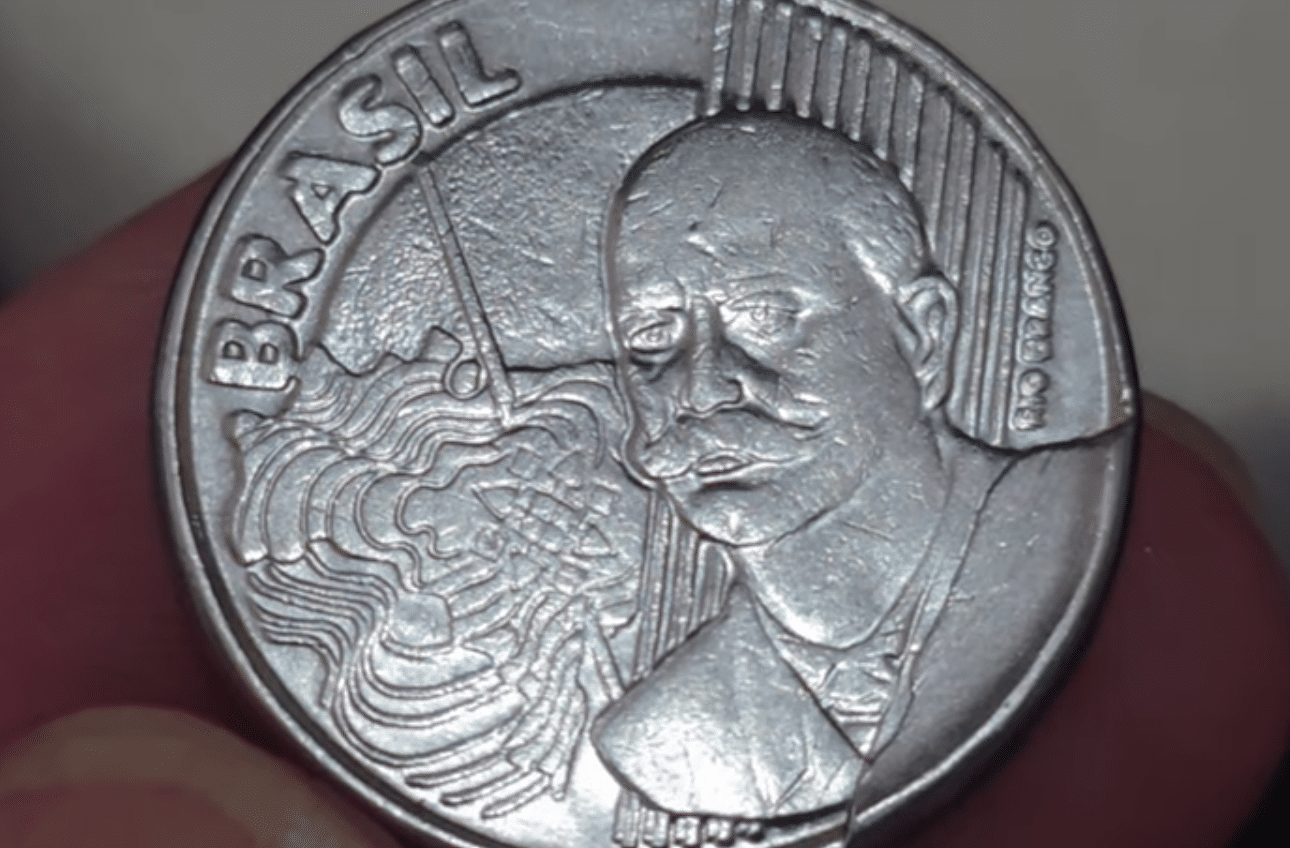 Imagem em close-up da moeda brasileira de 50 centavos, ano 2002. A foto foca no detalhe do reverso para auxiliar na identificação de erros de fabricação como o cunho trincado, que a tornam valiosa.