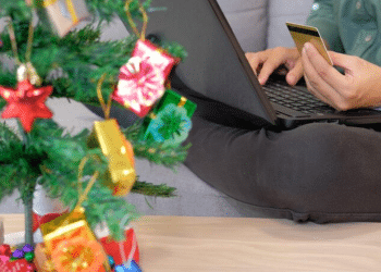 Pessoa realizando transação financeira pelo computador com cartão de crédito próximo a árvore de Natal decorada.