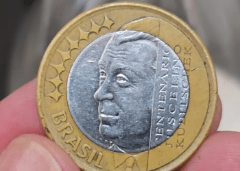 Mão segurando uma moeda de 1 real das Olimpíadas ou comemorativa do JK, mostrando detalhes do rosto de Juscelino Kubitschek e o anel dourado.