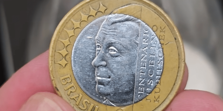 Mão segurando uma moeda de 1 real das Olimpíadas ou comemorativa do JK, mostrando detalhes do rosto de Juscelino Kubitschek e o anel dourado.