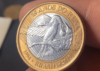 Moeda bimetálica de 1 real de 2019 sendo segurada pelos dedos, mostrando o reverso com a gravação de um beija-flor no ninho e a inscrição "25 Anos do Real".