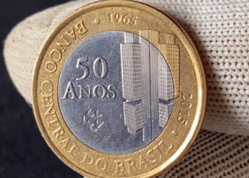 Close de uma mão usando luva branca segurando uma moeda de 1 real de 2015, comemorativa dos 50 anos do Banco Central, exibindo o núcleo prateado e o anel dourado.