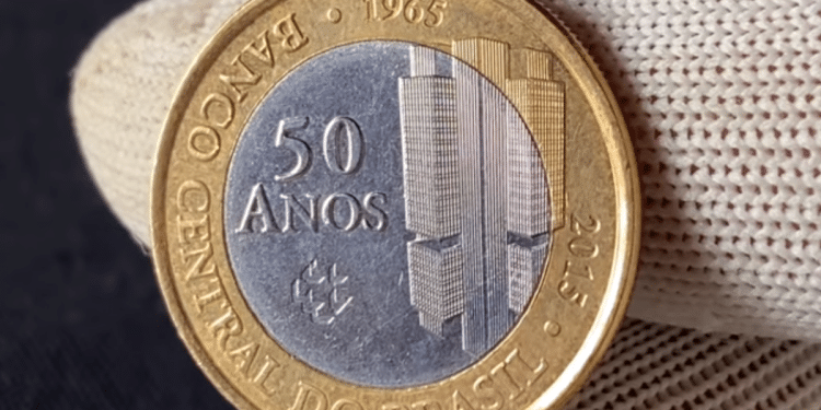 Close de uma mão usando luva branca segurando uma moeda de 1 real de 2015, comemorativa dos 50 anos do Banco Central, exibindo o núcleo prateado e o anel dourado.