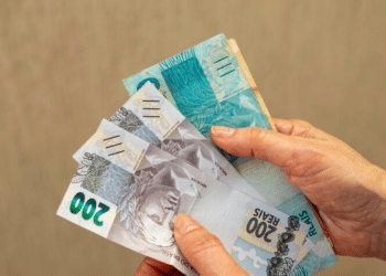 Duas mãos segurando um leque de notas de Real (100 e 200 reais) sobre um fundo cinza, demonstrando cédulas em bom estado de conservação.