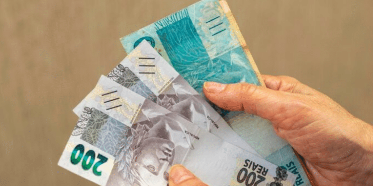 Duas mãos segurando um leque de notas de Real (100 e 200 reais) sobre um fundo cinza, demonstrando cédulas em bom estado de conservação.