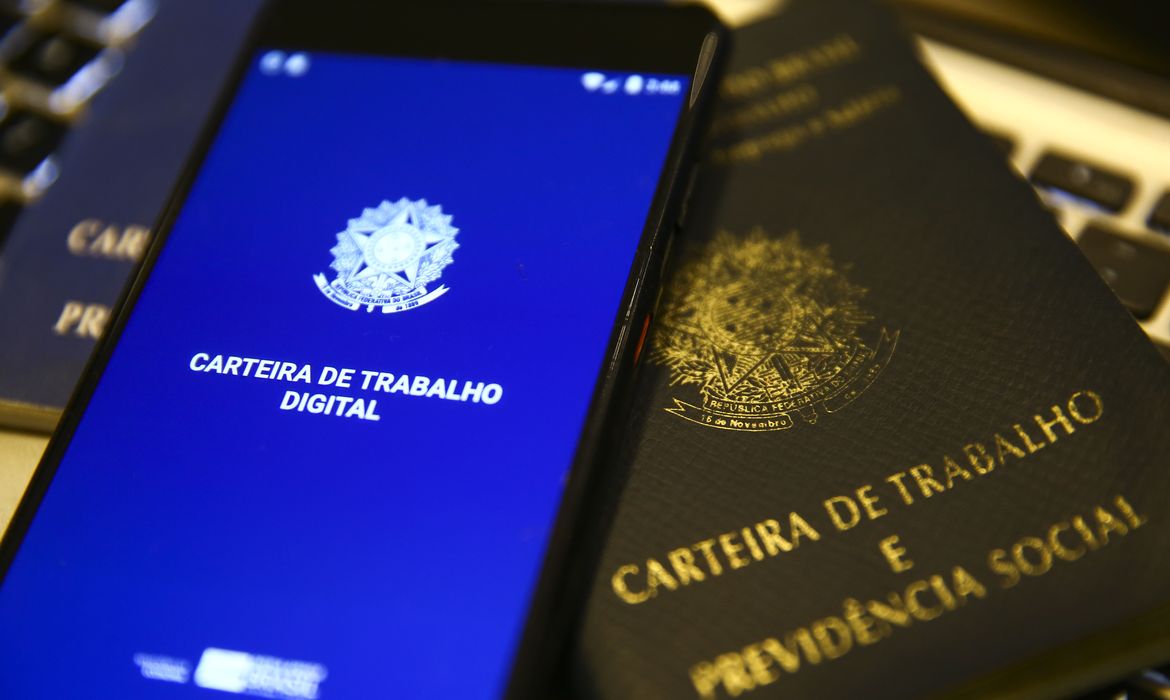 Smartphone exibindo Carteira de Trabalho Digital ao lado de carteira física sobre teclado de computador