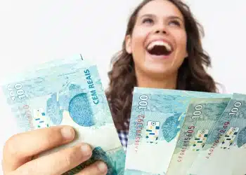 Pessoa feliz segurando notas de R$ 100, com sorriso no rosto, representando a liberação do auxílio de R$ 200 pelo governo entre 22 e 30 de dezembro.