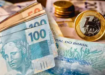 Composição com notas de 100 e 50 reais em primeiro plano, moedas empilhadas ao fundo e uma moeda de 1 real em destaque à direita.