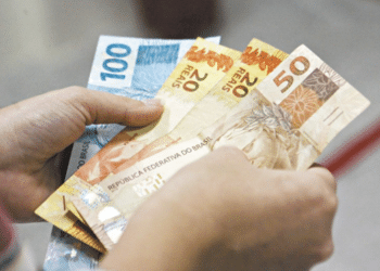 Duas mãos segurando notas de dinheiro em reais, incluindo cédulas de 100, 50 e 20 reais.