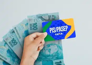 Mão segurando cartão PIS/PASEP da Caixa em frente a várias cédulas de 100 reais