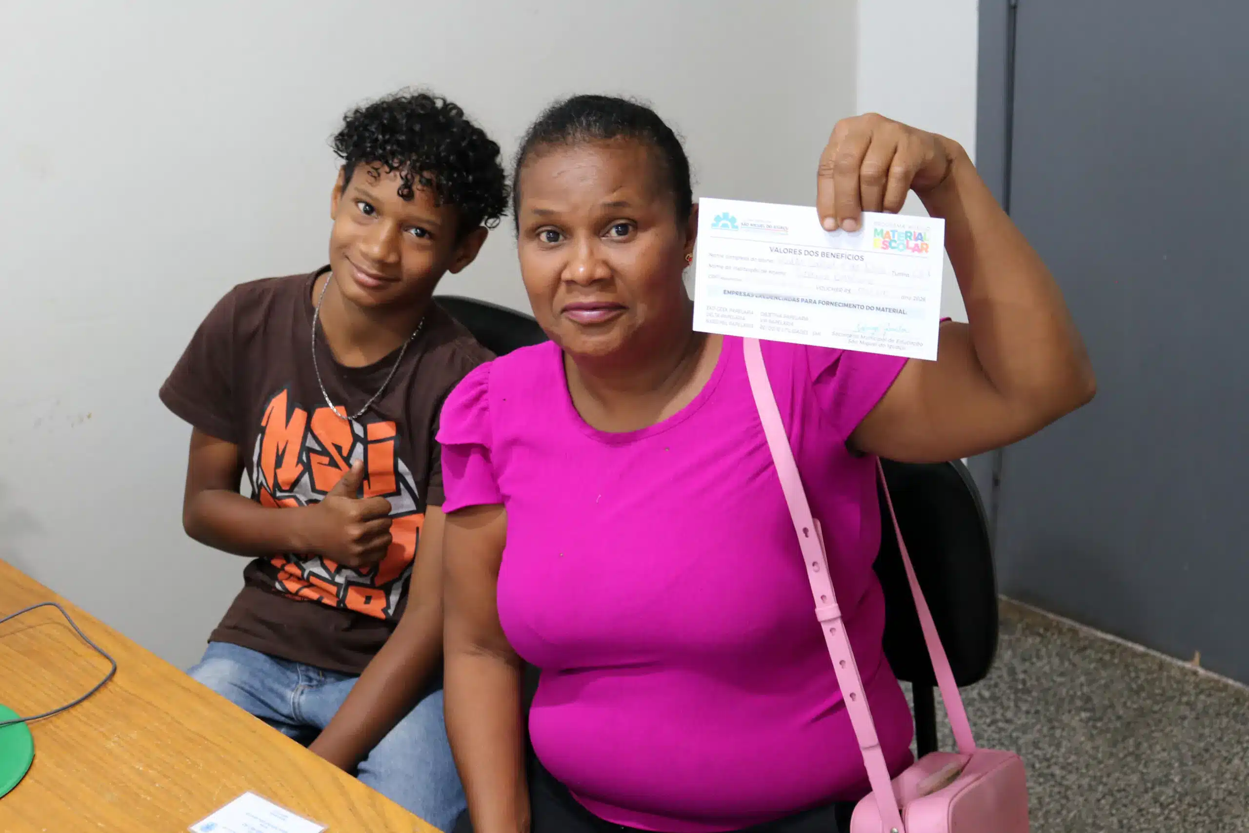 Auxílio Material Escolar 2026 Mulher segurando voucher do programa material escolar ao lado de menino jovem sorridente com camiseta preta.