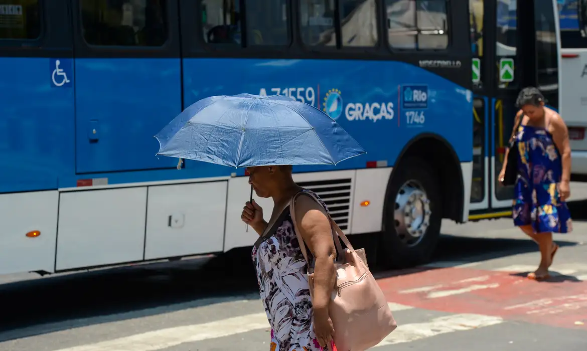 Clima 2026 Mulher caminhando na rua com sombrinha azul enquanto ônibus azul passa ao fundo no Rio de Janeiro