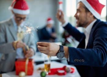 Pessoas vestindo chapéus de Papai Noel em festa de escritório, segurando uma vela de artifício e embalando presentes