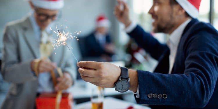 Pessoas vestindo chapéus de Papai Noel em festa de escritório, segurando uma vela de artifício e embalando presentes