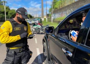 Agente de trânsito com colete e máscara verifica documentos apresentados por motorista em carro preto na via.