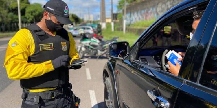 Agente de trânsito com colete e máscara verifica documentos apresentados por motorista em carro preto na via.