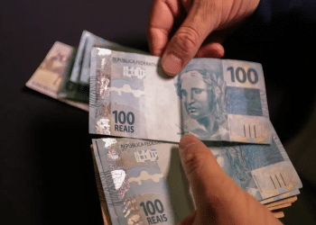 Mãos contando cédulas de 100 reais com fundo escuro desfocado.