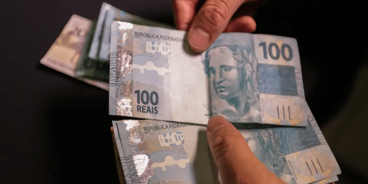 Mãos contando cédulas de 100 reais com fundo escuro desfocado.