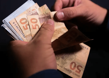 Mãos segurando e contando várias cédulas de cinquenta reais em fundo escuro.