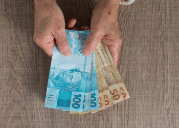 Mãos de pessoa idosa segurando notas de 100 e 50 reais sobre mesa de madeira.